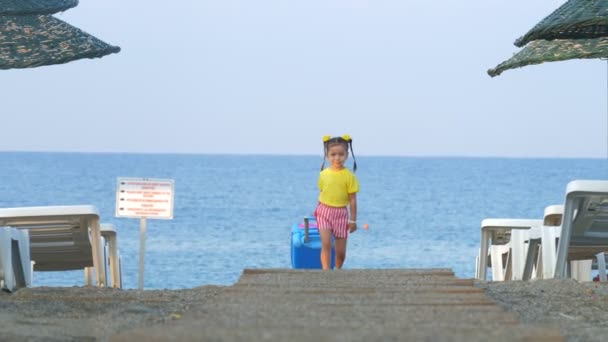 Klein schattig lachend meisje is wandelen met een koffer op een resort strand. — Stockvideo