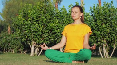 Kız, ağaçların yanındaki çimlere oturup yoga pozu veriyor..