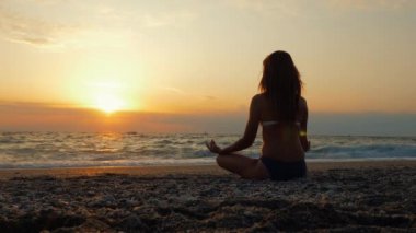 Bir genç kadın gün batımında deniz kenarında ya da okyanusta yoga yapıyor..