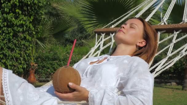 Mujer joven se encuentra en una hamaca con coctel de agua de coco . — Vídeos de Stock