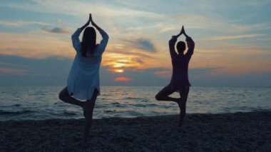 İki genç kadın gün batımında deniz kenarında ya da okyanusta yoga yapıyor..
