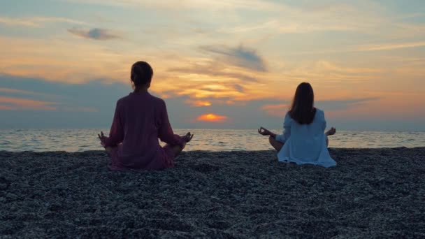 Dos mujeres jóvenes practican yoga junto al mar o el océano al atardecer . — Vídeo de stock