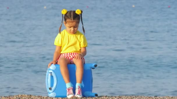 Kleines nettes Mädchen in bunten Kleidern sitzt auf einem blauen Koffer am Meeresstrand. — Stockvideo