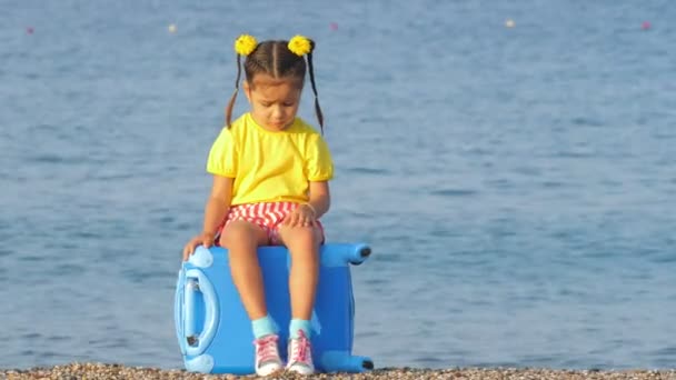 Niña linda en ropa colorida sentada en una maleta azul en la playa del mar . — Vídeo de stock