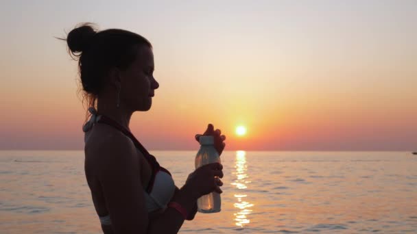Girl drinks water by the sea or ocean shore at sunset. — ストック動画