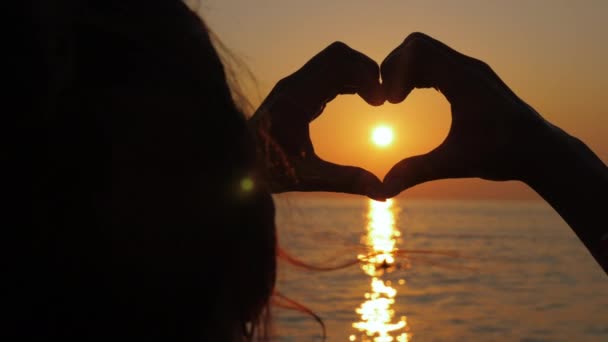 Mädchen hält Hände in Herzform auf dem Hintergrund der Sonne bei Sonnenuntergang. — Stockvideo