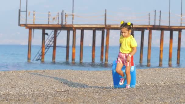Lilla söta leende flicka går med en resväska på en resort strand. — Stockvideo