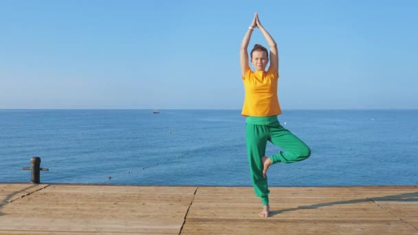 A young woman in colorful clothes practices yoga by the sea or ocean. — Stock Video