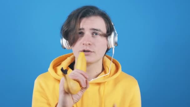 Ein junger Mann im Sweatshirt singt in einer Banane auf blauem Hintergrund. — Stockvideo