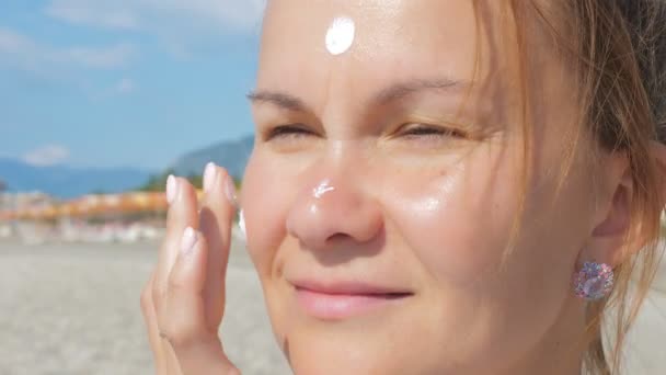Menina esfrega a pele do rosto com creme protetor solar enquanto está em pé em uma praia de areia . — Vídeo de Stock