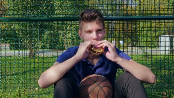 Feliz adolescente comiendo hamburguesa sentado en el campo de deportes — Vídeos de Stock