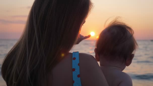 Mother holds baby in her arms while looking at sunset by the sea or ocean shore. — Stock Video