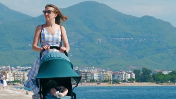 Madre joven camina a lo largo del paseo con un bebé en un cochecito . — Vídeos de Stock