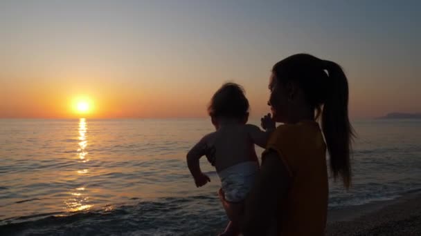 Mother holds baby in her arms while looking at sunset by the sea or ocean shore. — Stock Video
