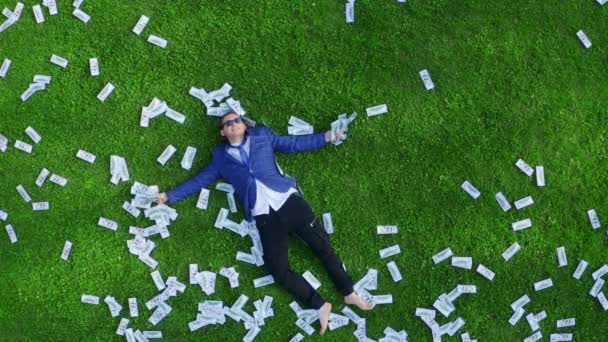 Young man businessman or student or office worker lies on a green grassy lawn strewn with money. — Αρχείο Βίντεο