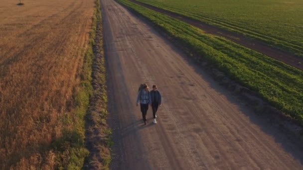 Hitchhikers and travelers, a young man with a girl, walk with backpacks on their shoulders along cereal fields sown with rye and wheat. — Stockvideo