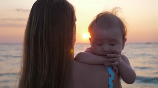 Anya a karjaiban tartja a babát, miközben a naplementét nézi a tenger vagy az óceán partján.. — Stock videók