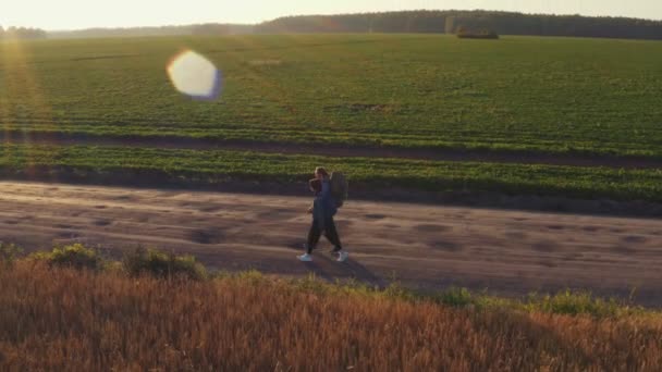 Autoestopistas y viajeros, un joven con una chica, caminan con mochilas sobre sus hombros a lo largo de campos de cereales sembrados con centeno y trigo . — Vídeos de Stock