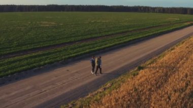 Hitchhikers and travelers, a young man with a girl, walk with backpacks on their shoulders along cereal fields sown with rye and wheat.