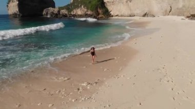 Deniz kayaları ve turkuaz okyanus, mavi gökyüzü ile plajda yürüyen bikinili Kadın geri görünümü. Atuh plajı, Nusa Penida adası, Bali, Endonezya. Tropikal arka plan ve seyahat konsepti. Drone Fotoğraf