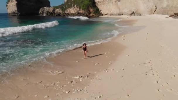 Вид сзади на женщину в бикини, идущую по пляжу с морскими камнями и бирюзовым океаном, голубым небом. Пляж Атух, остров Нуса Пенида, Бали, Индонезия. Тропический фон и концепция путешествия. Фотография дрона — стоковое видео