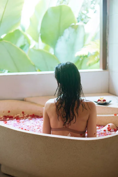 Rückansicht einer jungen Frau, die sich im Steinbad mit tropischen Blumen mit Blick auf den Dschungel entspannt, Bio-Hautpflege, luxuriöses Wellnesshotel, Lifestyle-Foto. Schönheitsbehandlungskonzept. — Stockfoto