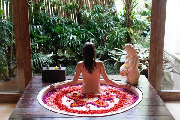 Vista posterior de morena Mujer joven en baño con pétalos de flores tropicales y aceites aromáticos. Tratamientos spa para el rejuvenecimiento de la piel. Chica relajante en bañera redonda de lujo —  Fotos de Stock