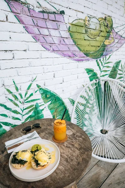 Hälsosam frukost med bröd och pocherat ägg med spenat, avokado och apelsinsmoothie på träbord.Modern design graffiti på en tegelvägg med avokado i en hängmatta — Stockfoto
