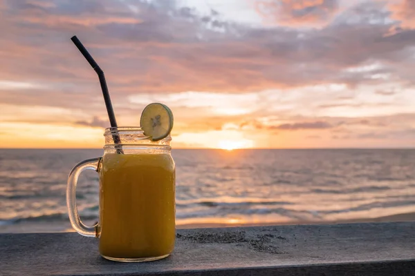 Glas mit bunten orangefarbenen Frucht Shakes auf dem Hintergrund des tropischen Strandes bei schönem rosa Sonnenuntergang mit Meerblick am Abend. Sommerstimmung. kalte Mixgetränke, Mango-Frucht-Smoothie. Raum für Text — Stockfoto