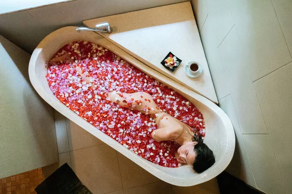돌로 된 욕조에서 쉬고 있는 여성의 맨 위의 모습붉은 색 꽃이 피고 있는 열 대 꽃 꽃잎, 유기농 피부 관리, 고급 온천 호텔, 생활 방식 사진 — 스톡 사진
