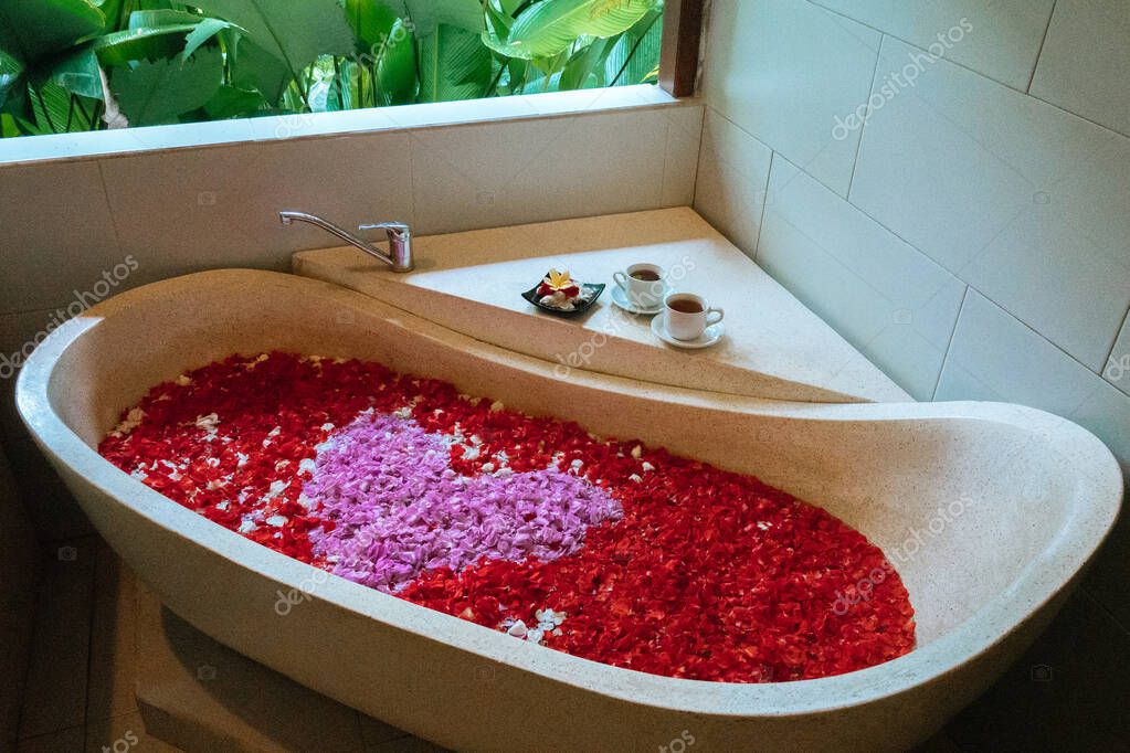 Vasca da bagno in pietra con petali di fiori a forma di cuore vicino alla  finestra con vista sulla giungla. Relax spa biologica nel lussuoso bagno di  Bali . - Foto Stock