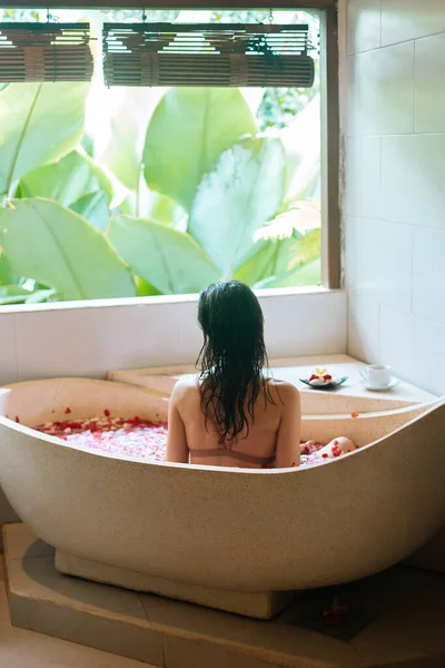 Widok z tyłu młodej kobiety kąpiącej się w kąpieli kwiatowej spa pełnej płatków. Luksusowy dzień spa z widokiem na dżunglę. Koncepcja dobrego samopoczucia, pielęgnacji ciała i pielęgnacji ciała — Zdjęcie stockowe