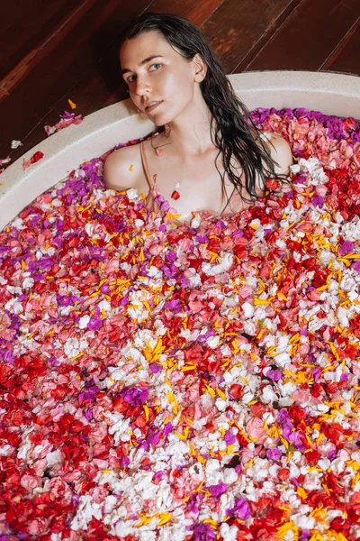 Hermosa joven disfrutando en el spa al aire libre. Lujosa bañera redonda. Pétalos tropicales orgánicos naturales en el agua: blanco, rosa, rojo, violeta. Concepto de tratamiento de belleza . —  Fotos de Stock