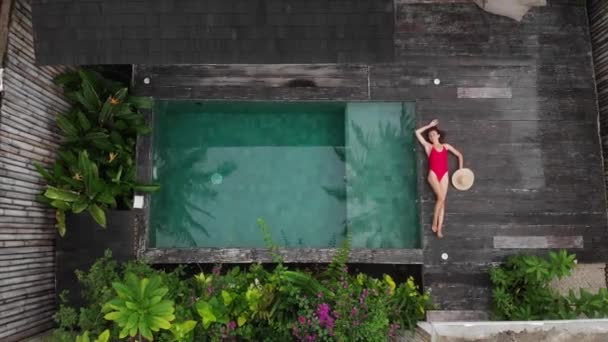 Vista dall'alto della felice Donna in costume da bagno rosso e cappello di paglia prendere il sole vicino alla piscina privata con fiori e verde intorno, Bali. Contesto tropicale e concetto di viaggio . — Video Stock