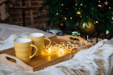 Sıcak Noel kompozisyonu Tahta bir tepside sıcak içecekler ile iki sarı kupa yatak üzerinde pofuduk bir battaniye ile arkaplanda Noel ağacı dekor iç oda çelenk