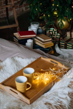 Sıcak Noel kompozisyonu Tahta bir tepside sıcak içecekler ile iki sarı kupa yatak üzerinde pofuduk bir battaniye ile arkaplanda Noel ağacı dekor iç oda çelenk