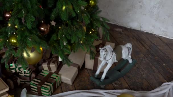 Vacaciones, cajas de regalo de Año Nuevo y juguete de caballo de madera bajo el árbol de Navidad decorado con bolas de oro, guirnalda en el acogedor interior del loft . — Vídeo de stock