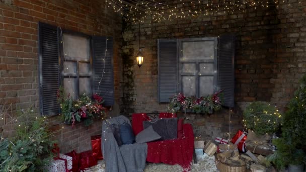 Szilveszter és mágia díszített külseje karácsonyfa, sok ajándék és ünnepi fények és koszorúk házon kívül kanapéval és kockás háttér téglafal. — Stock videók