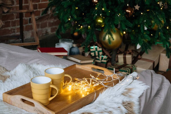 Cozy Christmas composition, two yellow mug with hot drinks on a wooden tray stand on the bed with a fluffy blanket against the background of Christmas tree in loft interior room in decor in garlands