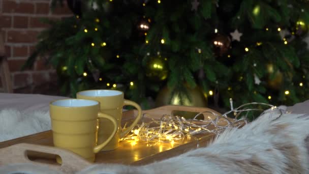 Mysig jul sammansättning, två gula mugg med varma drycker på en trälåda stå på sängen med en fluffig filt mot bakgrund av julgran i loft interiör rum i inredning i girlanger — Stockvideo
