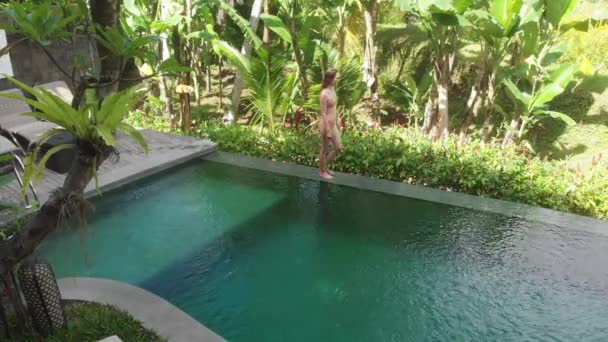 Luftaufnahme des Infinity-Pools auf der Villa mit tropischem Garten. Frau, die am Rande des Pools spaziert und in beigem Bikini und Hut die Aussicht auf den Dschungel genießt. Urlaubskonzept — Stockvideo