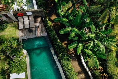 Tanınmayacak kadar zayıf, bej bikinili genç bir kadının Ubud.Tatil konsepti içinde yeşil tropik bir havuzda güneşlenmesidir. Drone fotoğrafı