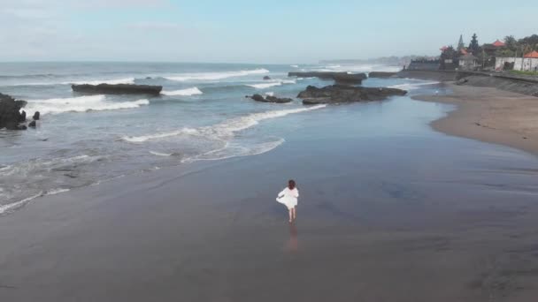 Вид на щасливу жінку в білій сукні біжить по береговій лінії і знімає відео з дроном на пляжі з чорним піском. Відпустка на Балі. — стокове відео