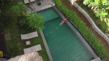 Kırmızı bikinili sıska bir kadının havadan görünüşü rahatlatıcı ve Ubud 'daki tropik bahçe villasında yüzme havuzunda güneşleniyor. Drone yükseliyor. Tatil kavramı.