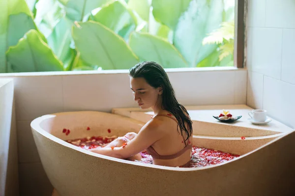 Back view of Woman relaxing in stone bath tube with tropical flowers with jungle view, organic skin care, luxury spa hotel, Bali Indonesia — 스톡 사진