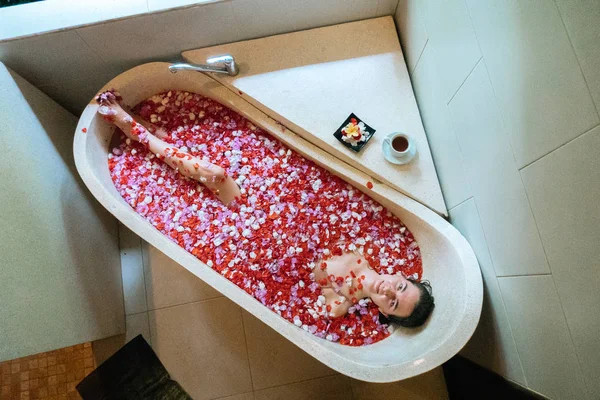 Vista superior de la mujer relajante en bañera de piedra con pétalos de flores tropicales en colores violeta y rojo, cuidado de la piel orgánica, hotel spa de lujo, foto estilo de vida —  Fotos de Stock