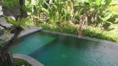 Tropik bahçeli villa üzerindeki Sonsuzluk Havuzu 'nun havası. Havuzun kenarında yürüyen ve orman manzarasından hoşlanan bir kadın bej bikini ve şapka giyiyor. Tatil kavramı