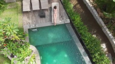 Havadan bej bikinili genç bir kadının rahatlama görüntüsü ve Ubud 'daki şezlonglu ormanda yüzme havuzunun yanında güneş banyosu yapması. Drone yükseliyor. Tatil kavramı.