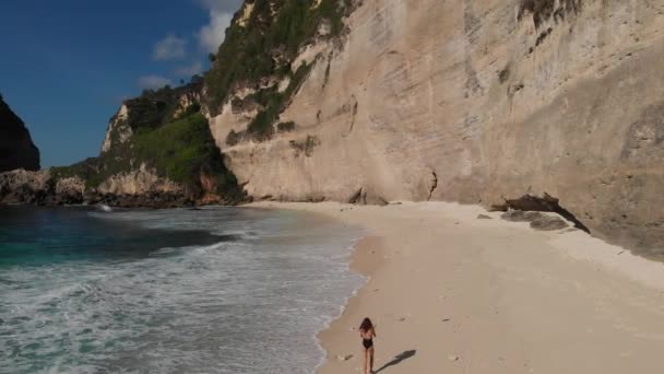 Вид сзади на женщину в бикини, идущую по пляжу с морскими камнями и бирюзовым океаном, голубым небом. Пляж Атух, остров Нуса Пенида, Бали, Индонезия. Тропический фон и концепция путешествия. Дрон — стоковое видео