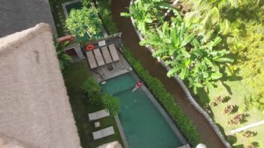 Kırmızı bikinili sıska bir kadının havadan görünüşü rahatlatıcı ve Ubud 'daki tropik bahçe villasında yüzme havuzunda güneşleniyor. Drone düşüyor. Tatil kavramı.